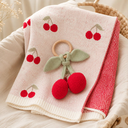 A folded baby blanket or throw in a woven basket, featuring a cream front with red cherry prints, a red textured reverse side, and a wooden ring with a knotted green bow and red pom-pom cherries attached.