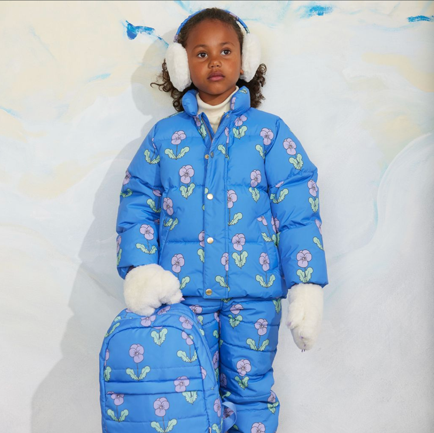 A young child wearing a bright blue puffer snowsuit with a floral print, matching hat, white earmuffs, and white gloves, posed against a light textured backdrop.