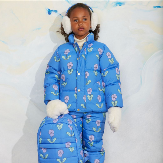 A young child wearing a bright blue puffer snowsuit with a floral print, matching hat, white earmuffs, and white gloves, posed against a light textured backdrop.