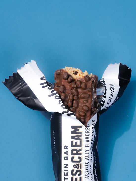 A partially unwrapped protein bar with chocolate-covered clusters and a beige cracker base, shown on a bright blue background. The wrapper is black and white with visible text including 'PROTEIN BAR' and 'CHOCOLATE & CREAM'.