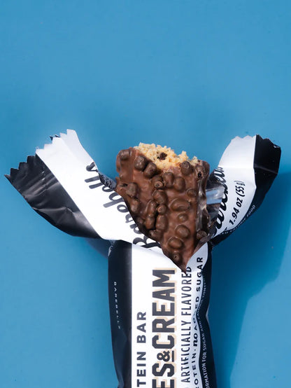 A partially unwrapped protein bar with chocolate-covered clusters and a beige cracker base, shown on a bright blue background. The wrapper is black and white with visible text including 'PROTEIN BAR' and 'CHOCOLATE & CREAM'.