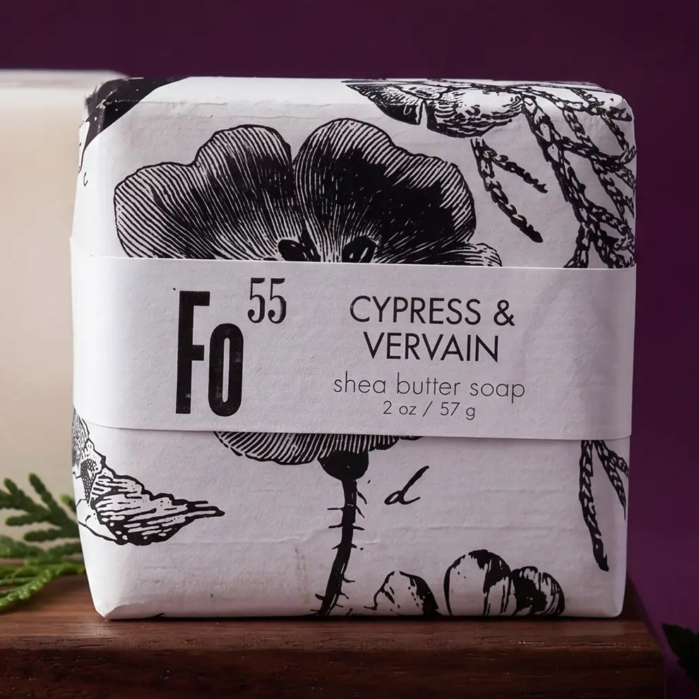 A wrapped bar of soap labeled 'Fo 55 Cypress & Vervain' sits on a wooden surface against a dark purple background. The soap is wrapped in white paper with black botanical illustrations and a white paper band around the middle.