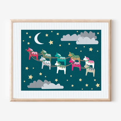 A framed art print featuring a whimsical night sky with a crescent moon, stars, clouds, and colorful Dala-style horses carrying a sack on a sled, shown against a light wall.