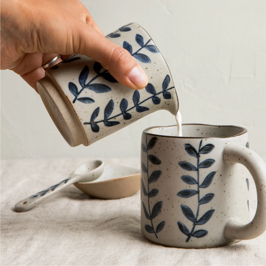 Person pouring liquid from one ceramic mug with leaf pattern into another on a neutral background