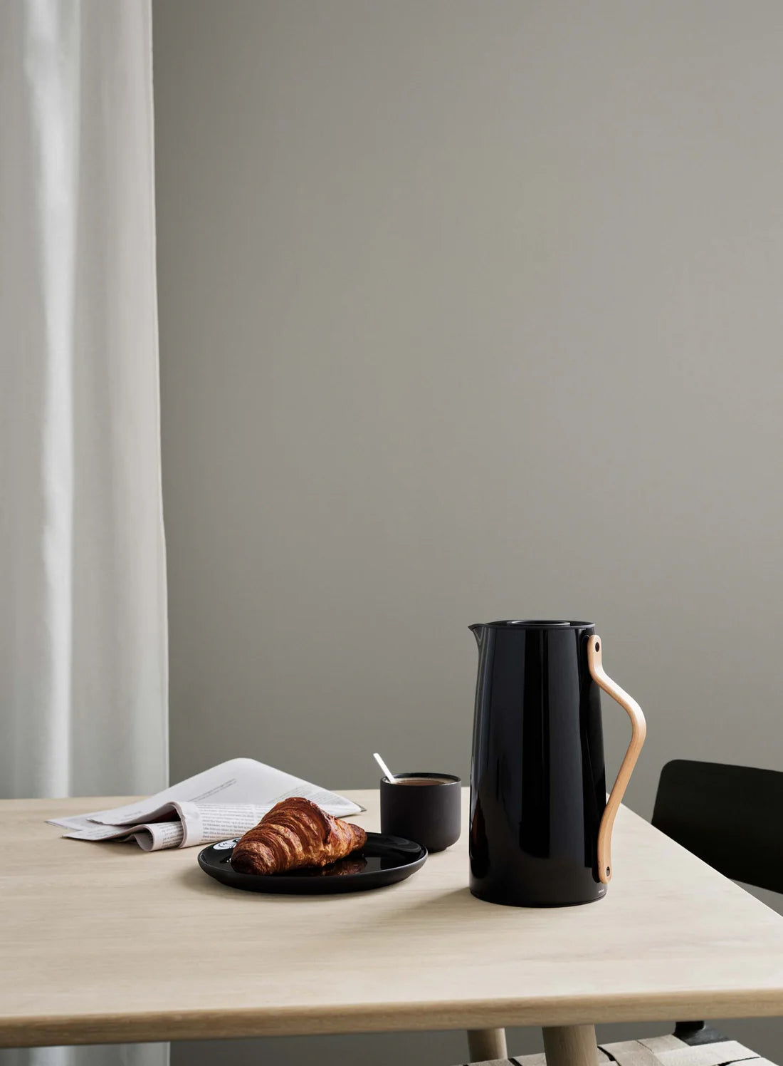 Black ceramic pitcher on a wooden table with a croissant and coffee cup against a gray wall.