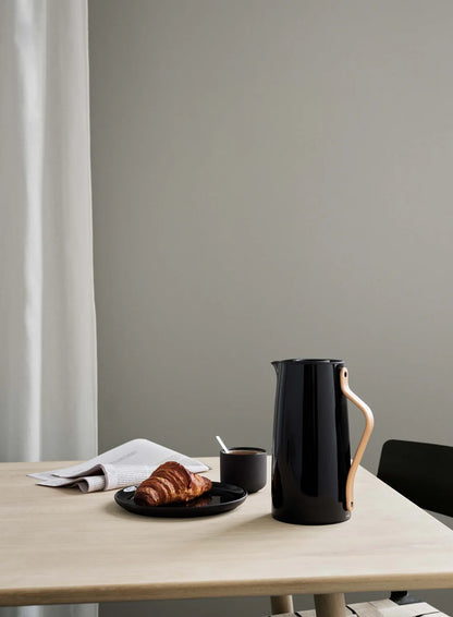 Black ceramic pitcher on a wooden table with a croissant and coffee cup against a gray wall.