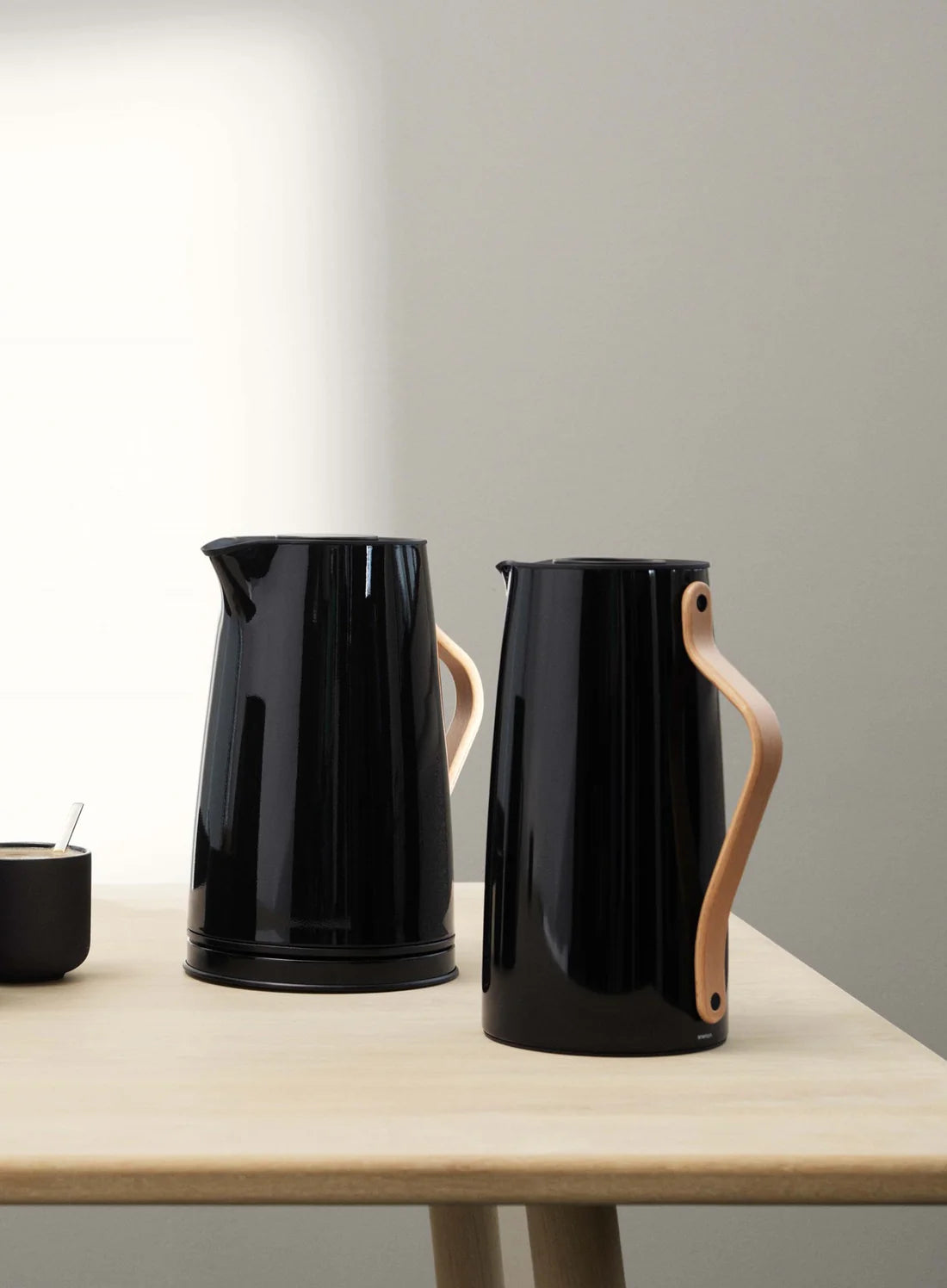 Two black ceramic pitchers with wooden handles on a light wooden table against a plain wall.