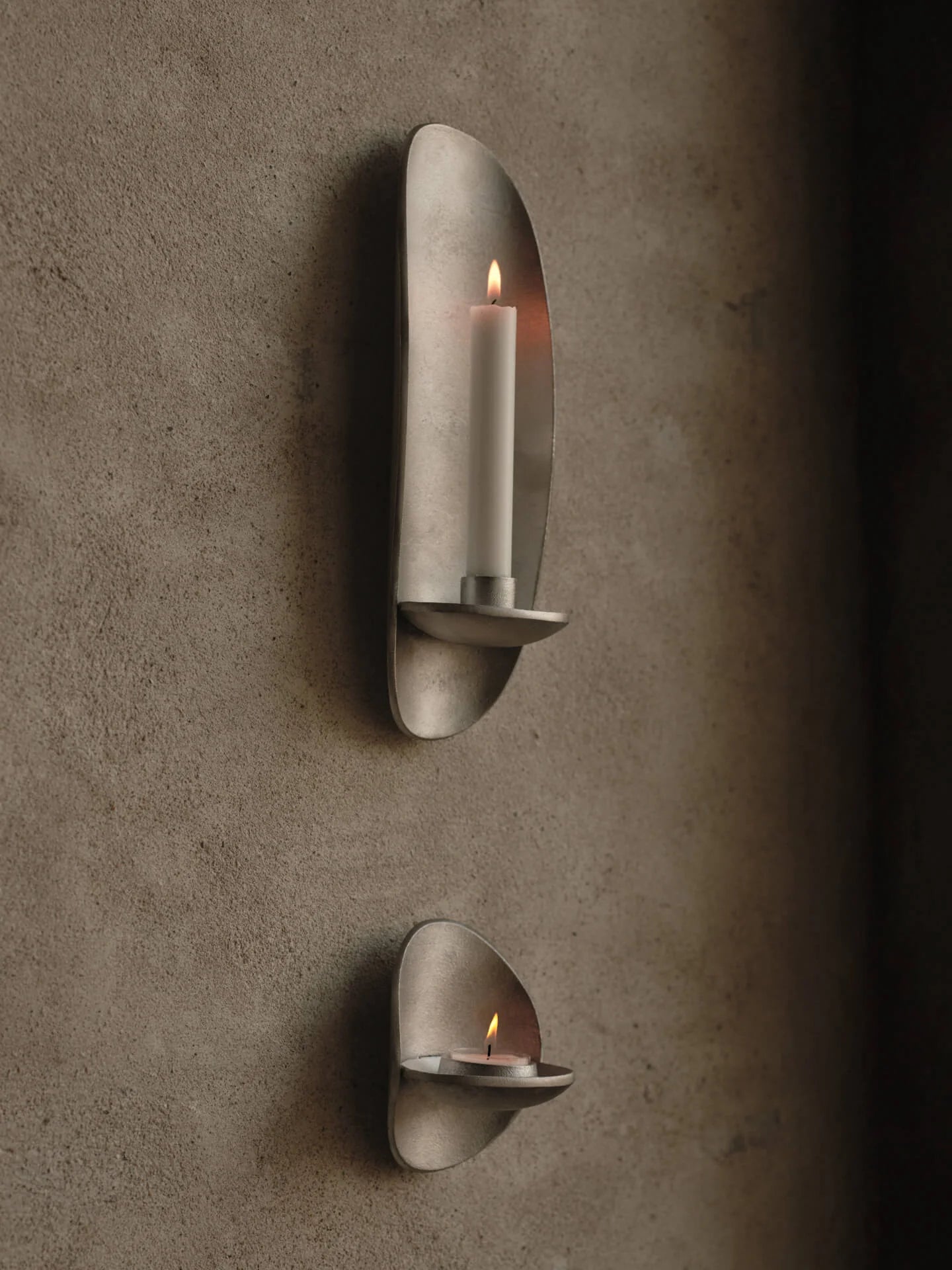 Two metallic wall-mounted candle holders with lit candles on a texturedNamed after the Greek word for ‘spiritual love and charity,’ the Agapé Wall Candle Holder is crafted from recycled cast aluminium. Its rustic surface creates a mesmerising ambience, infusing your surroundings with a sense of warmth and spirituality while adding a touch of elegance. Designed to fit a 2.2 cm taper candle, the Agapé Wall Candle Holder is wall-mounted, seamlessly combining functionality with timeless beauty. wall.
