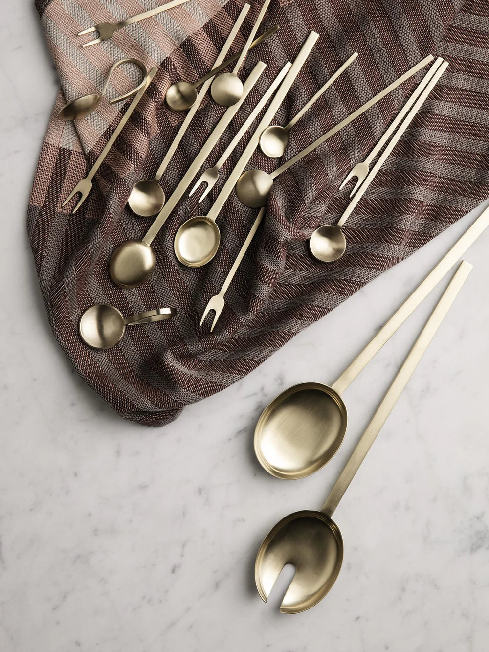 Set of gold-colored spoons and forks on a marble surface with a brown cloth underneath.