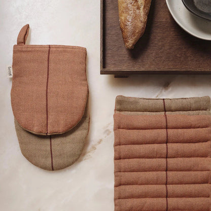 A set of oven mitts and a pot holder in a neutral color palette, displayed on a marble surface with a baguette and a cup in the background.