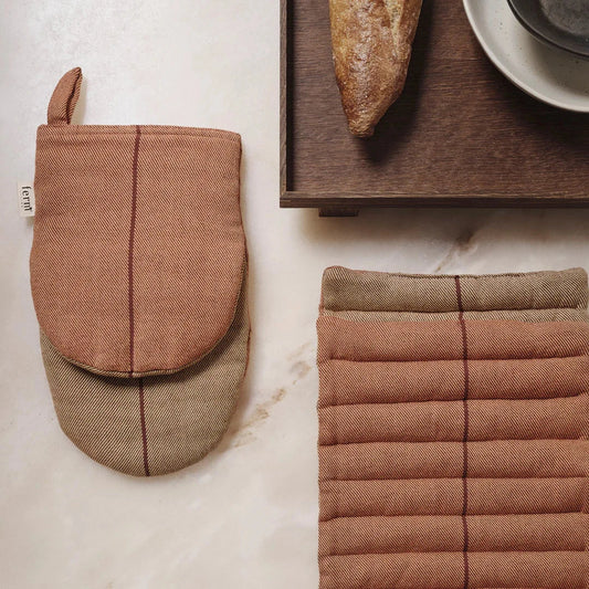 A set of oven mitts and a pot holder in a neutral color palette, displayed on a marble surface with a baguette and a cup in the background.