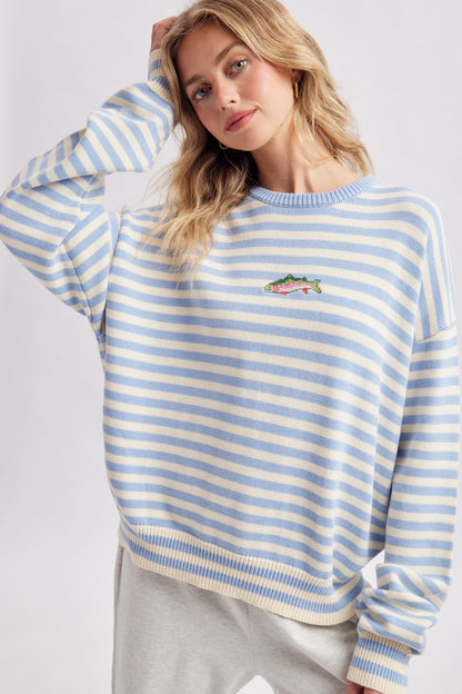 A woman wearing a light blue and white striped crewneck sweatshirt with a small embroidered fish on the left chest, paired with light gray sweatpants, against a plain light gray background.