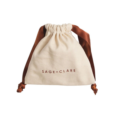 An off-white canvas drawstring pouch with brown satin drawstrings and the brand name 'SAGE+CLARE' printed in brown on the front, set against a plain white background.