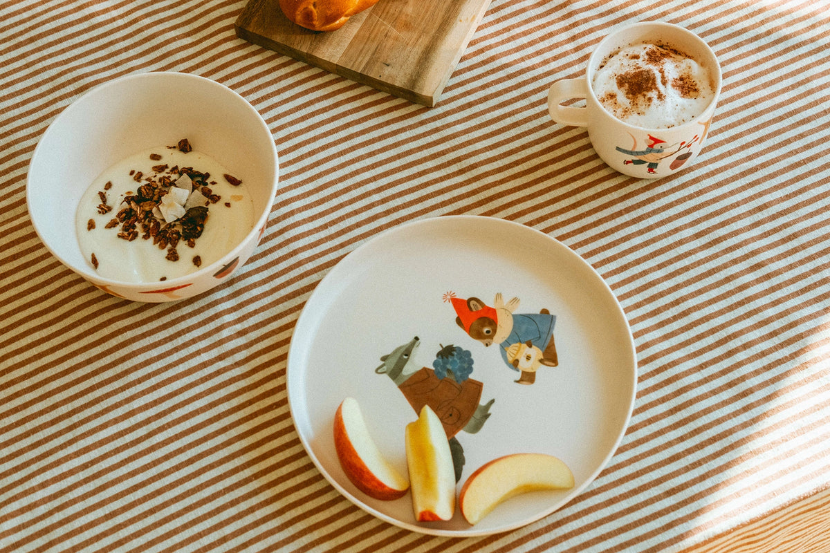 A styled tabletop scene with a white ceramic plate featuring a colorful cartoon forest illustration, containing apple slices, alongside a white bowl with granola and a white mug topped with whipped cream and cocoa powder, all arranged on a beige-and-white striped tablecloth.