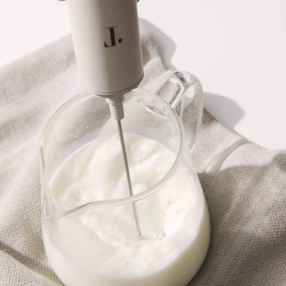 Handheld electric mixer with a clear glass container filled with whipped cream on a light fabric background.