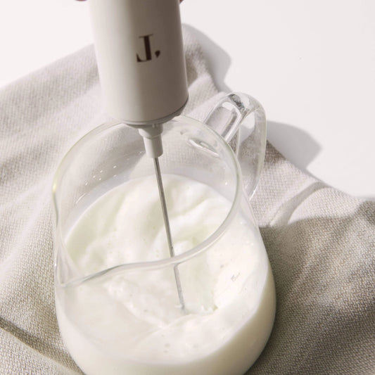 Handheld electric mixer with a clear glass container filled with whipped cream on a light fabric background.