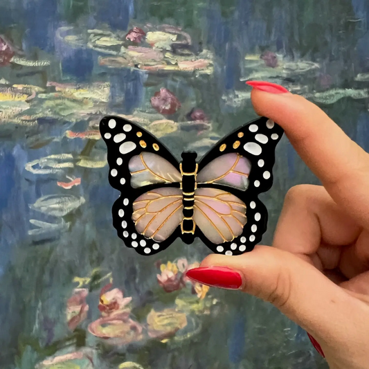 Butterfly-shaped object held by a hand with red nail polish against a blurred painting of water lilies.