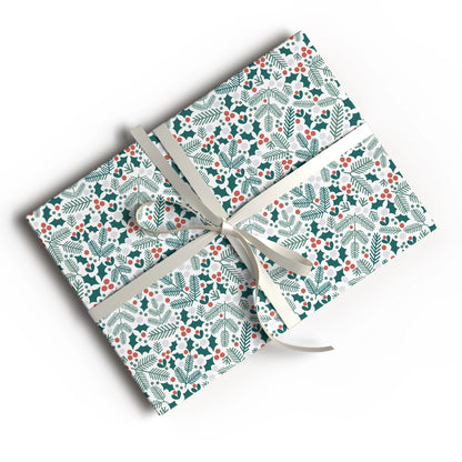 A gift box wrapped in festive holiday paper with a green and red holly and pine pattern, tied with a cream ribbon, shown on a white background.