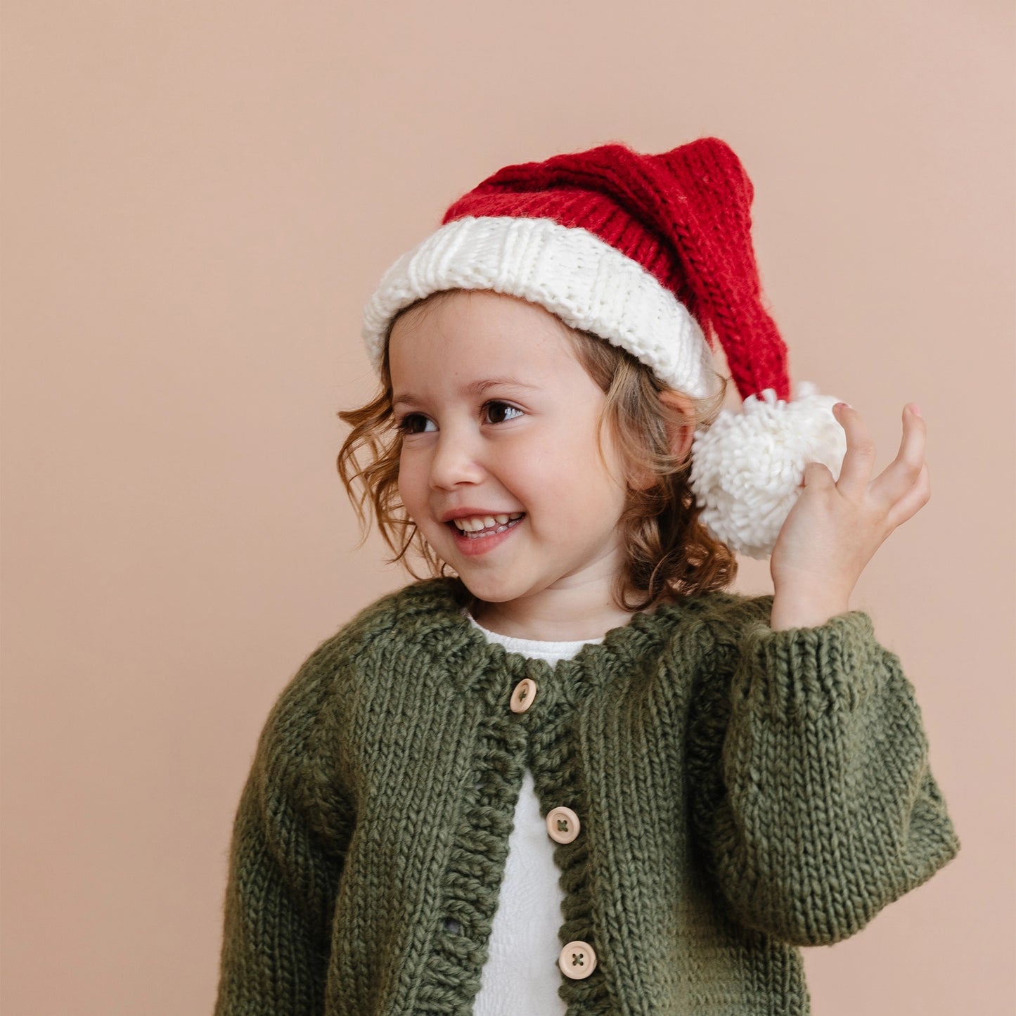 Child wearing a green knitted cardigan and red and white Santa hat on a beige background