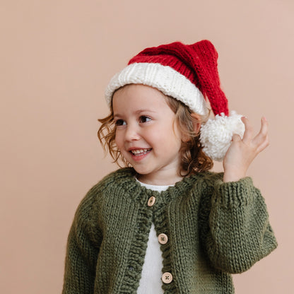 Child wearing a green knitted cardigan and red and white Santa hat on a beige background