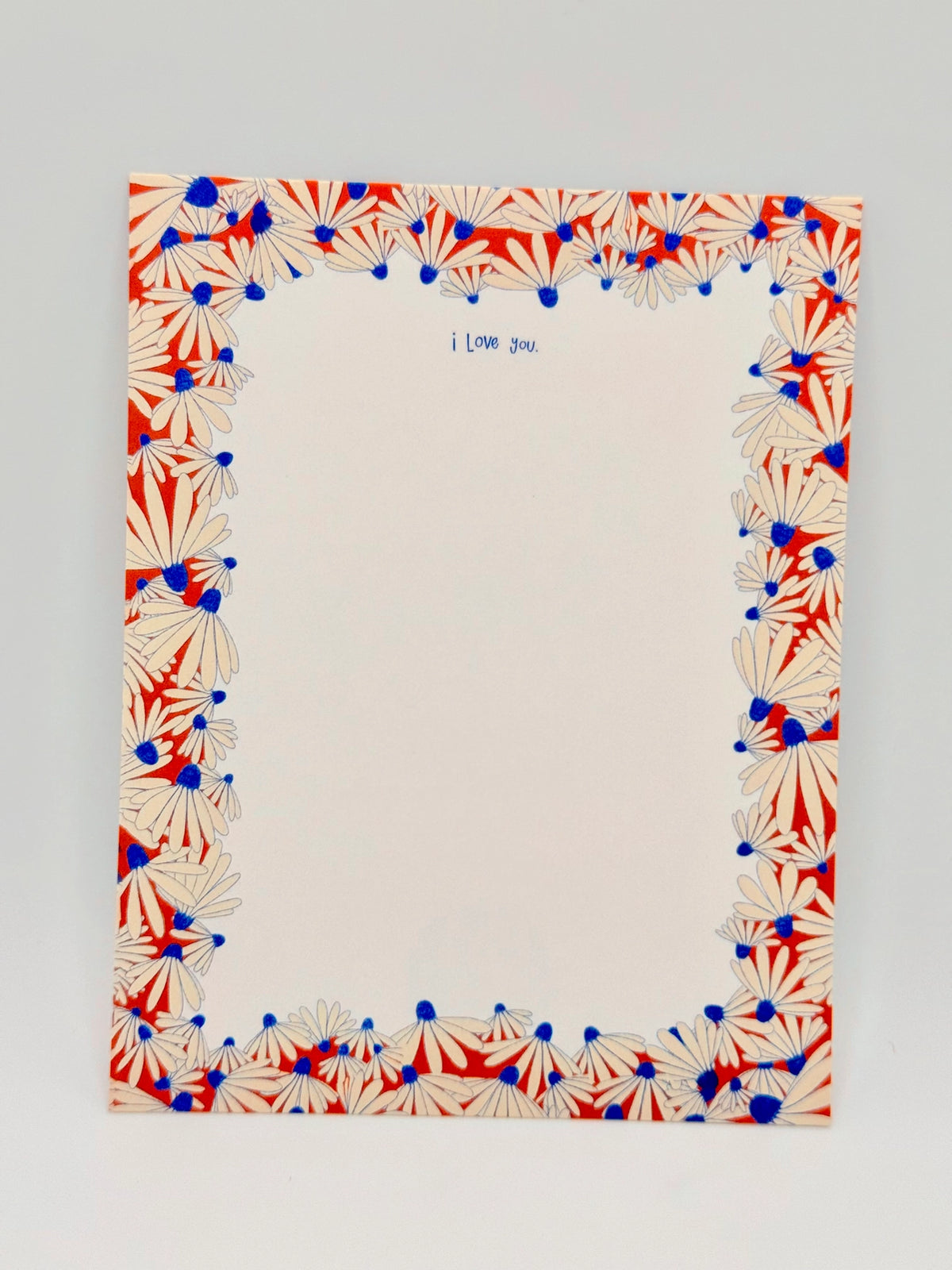 A square cream-colored card or note with a decorative border of red and blue daisy-like flowers around the edges, with the handwritten text 'I Love you' at the top center, shown on a light gray background.