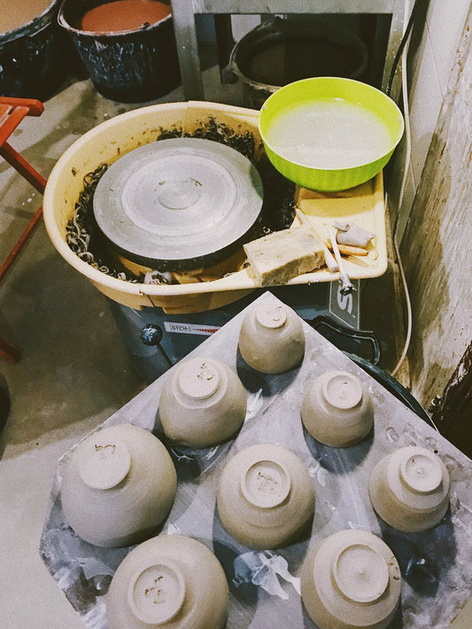 Several unglazed ceramic bowls are arranged on a plastic-covered tray in front of a pottery wheel, with a green bowl and other pottery tools visible in the workshop.