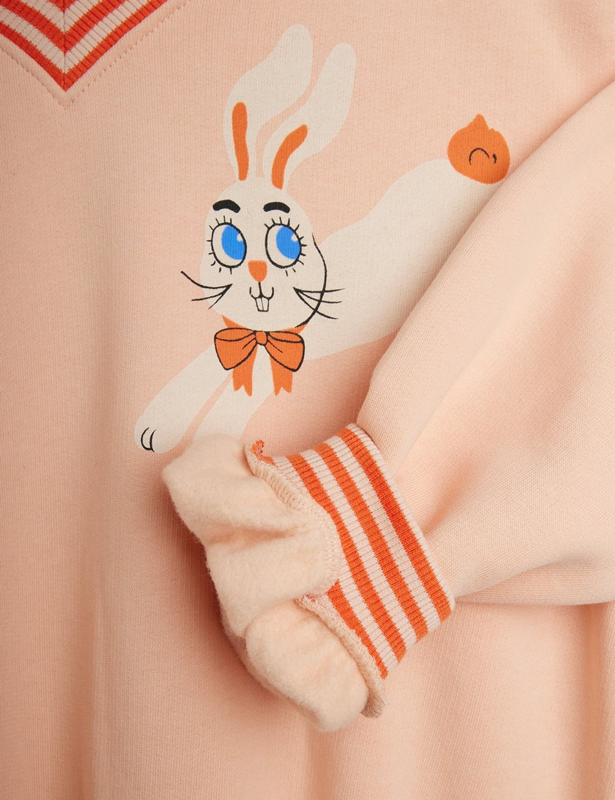 Close-up of a light peach-pink sweatshirt featuring a cartoon bunny graphic with blue eyes and an orange bow tie, plus orange-and-white striped ribbed cuffs.