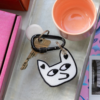 Keychain with a cat design on a surface next to a pink cup and pink box.
