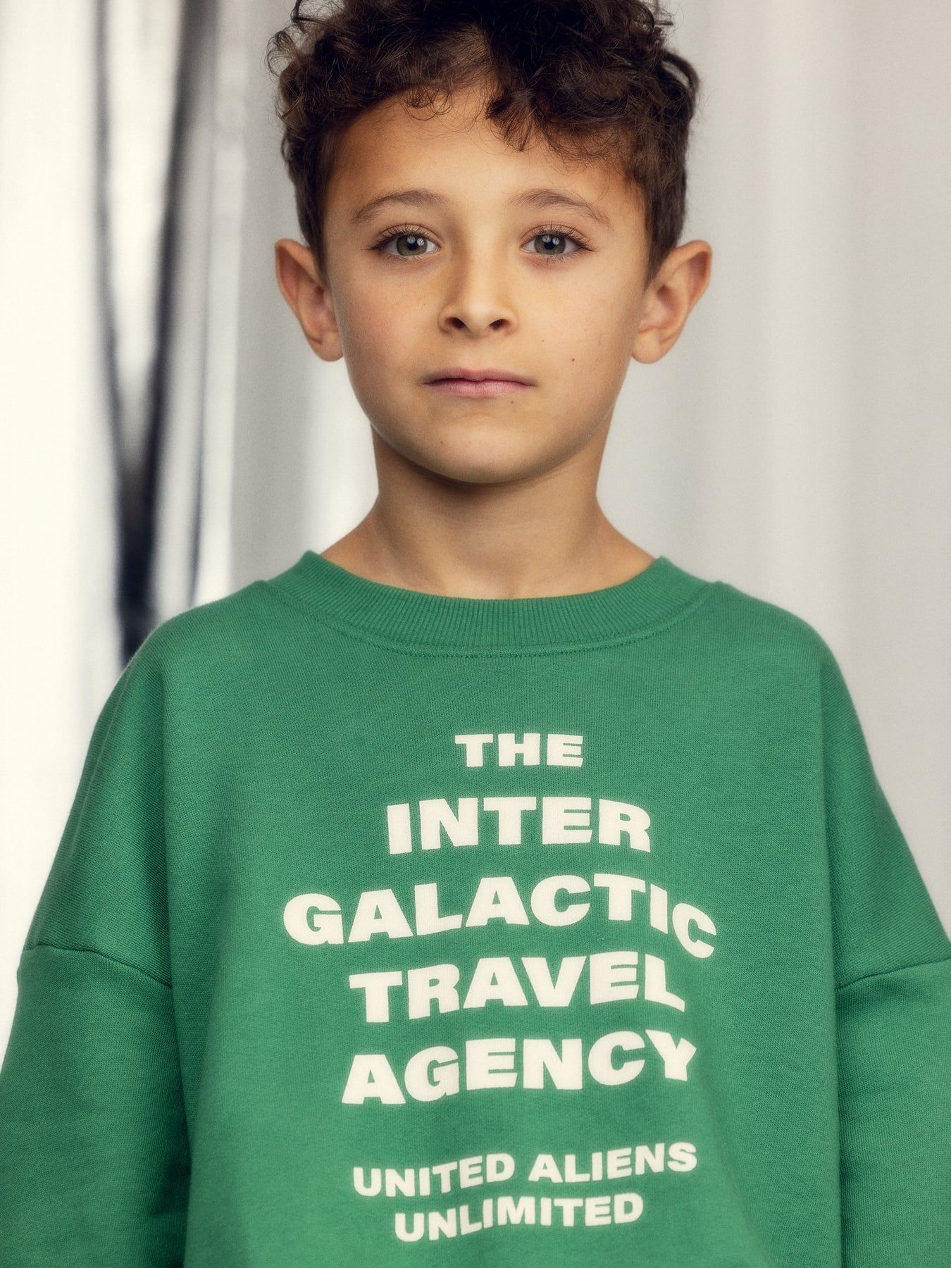 Child wearing a green sweatshirt with text 'The Inter Galactic Travel Agency United Aliens Unlimited' against a neutral background.