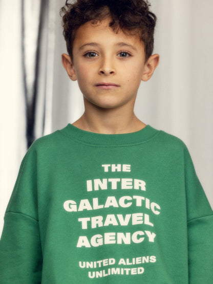 Child wearing a green sweatshirt with text 'The Inter Galactic Travel Agency United Aliens Unlimited' against a neutral background.