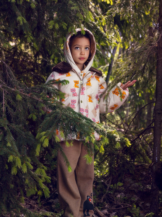 A young girl stands in a dense evergreen forest wearing a cream hooded jacket with a colorful pattern of cartoon animals and brown pants.