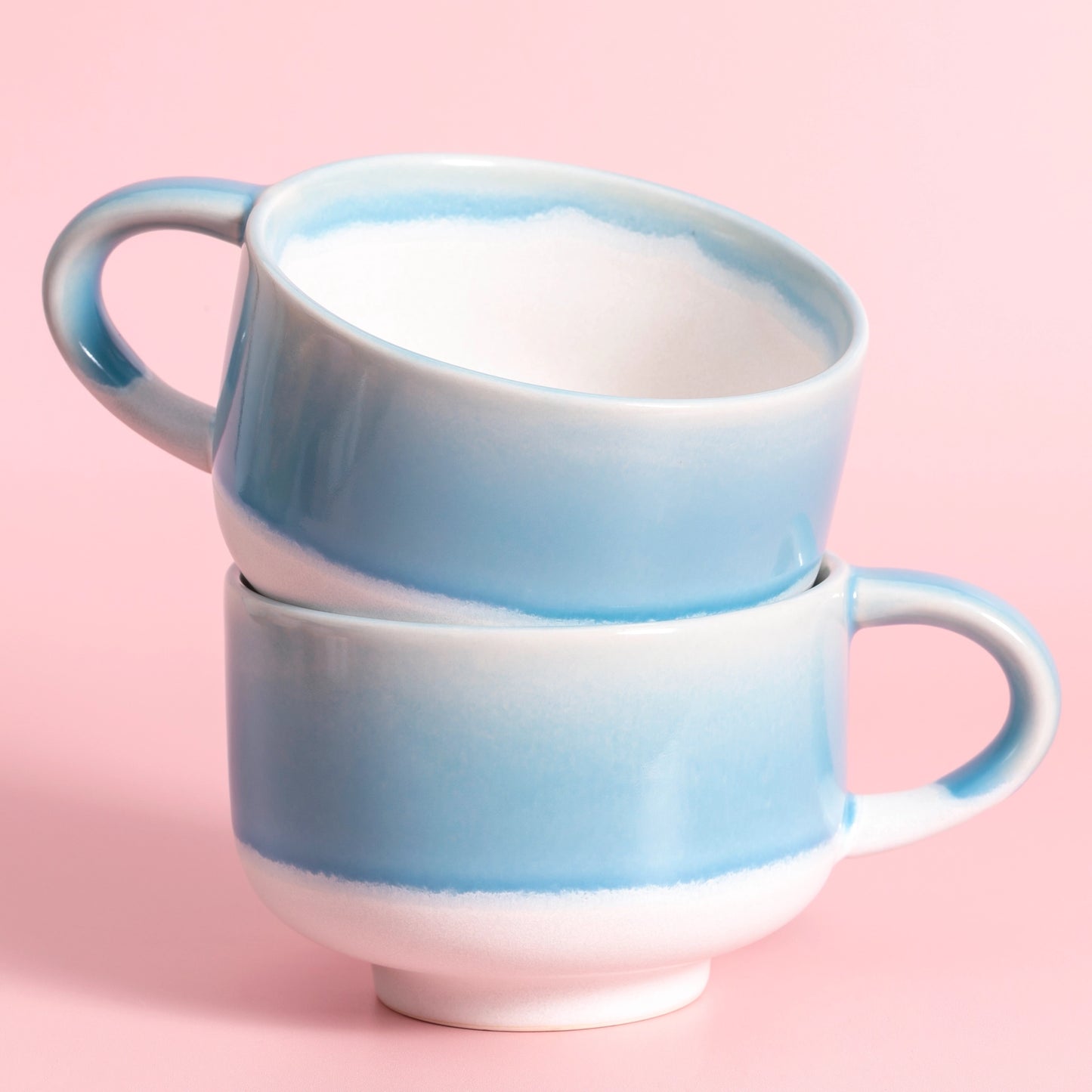 Two stacked ceramic mugs with a light blue and white gradient glaze, set against a solid pink background.