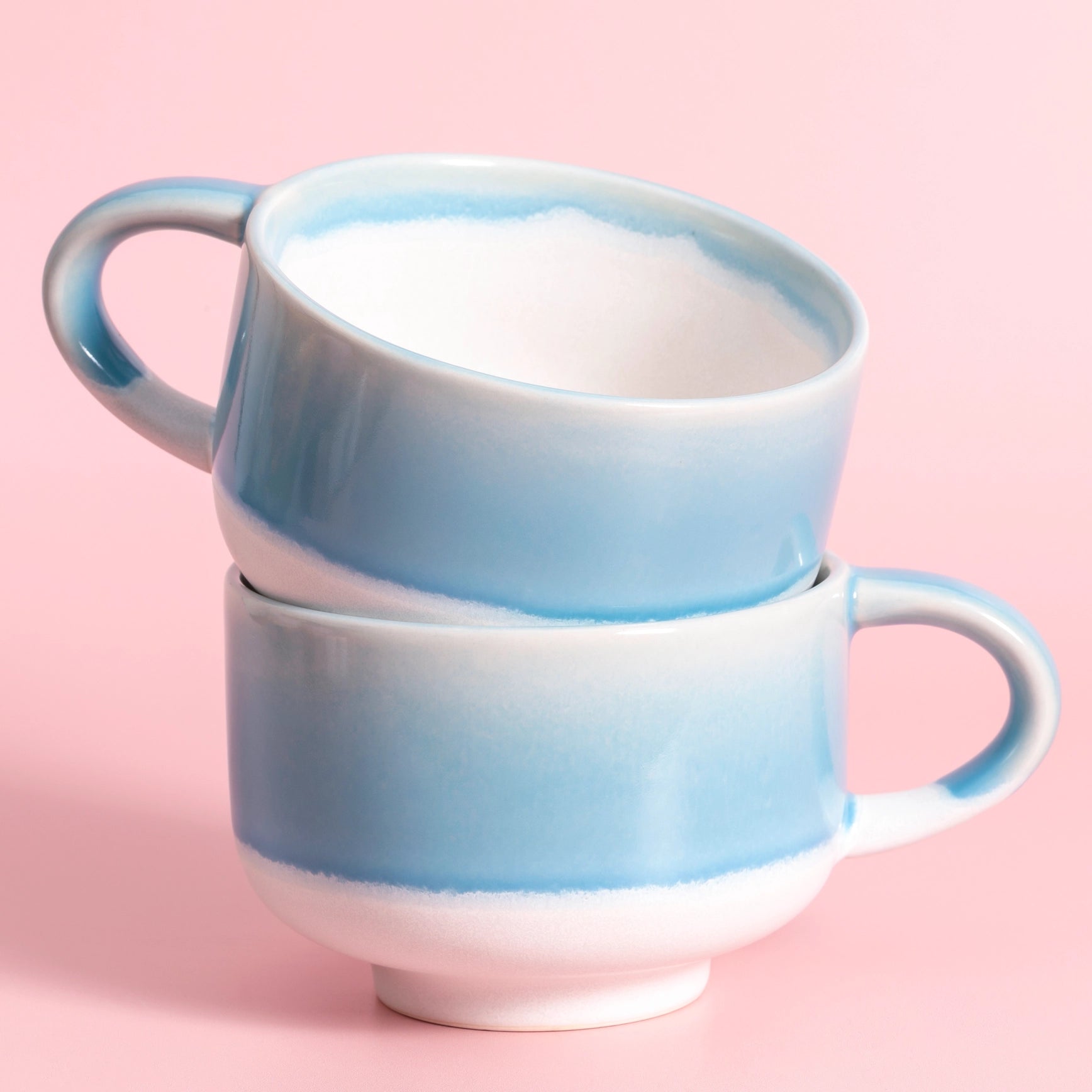 Two stacked ceramic mugs with a light blue and white gradient glaze, set against a solid pink background.