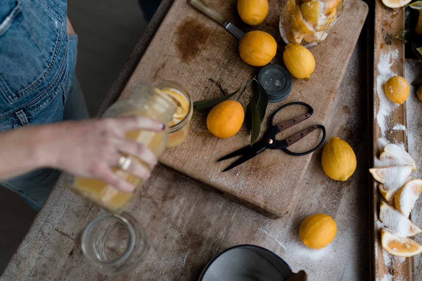 Person preparing lemons on a wooden cutting board with a jar and scissors.