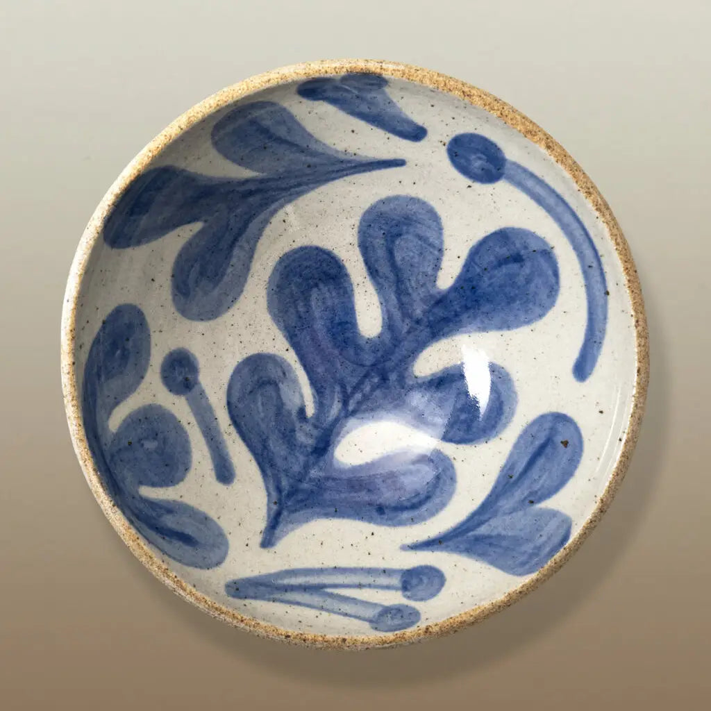 A handmade ceramic plate with a blue floral pattern on a speckled off-white glaze, featuring a rustic, unglazed beige rim.