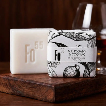Two bars of Fo 55 Shea Butter Soap displayed on a dark wooden block. The left bar is solid white with the Fo 55 logo embossed, and the right bar is wrapped in a white paper sleeve with black botanical illustrations and a label reading 'Mahogany & Cognac'.