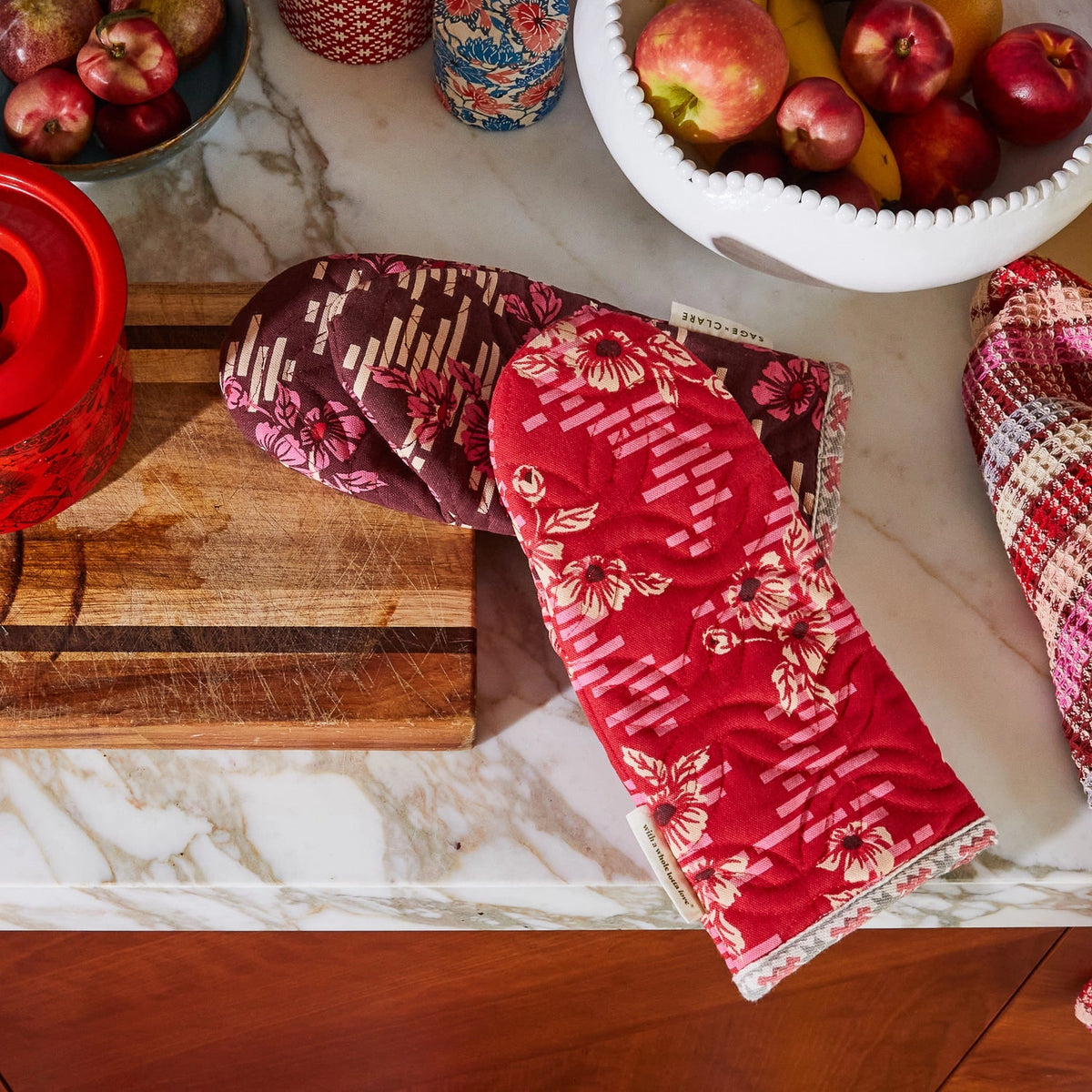 Red floral oven mitts on a marble countertop with apples and a cutting board.