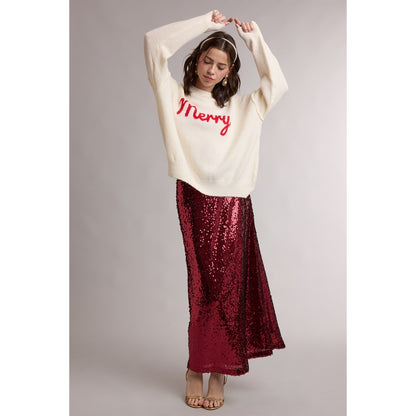 Woman with hands above her head wearing long sequin skirt with an of-white, holiday sweater with the word "Merry" written across it