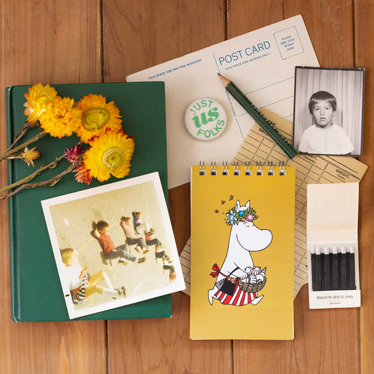 Collection of items including a notebook, postcard, and Moomin character book on a wooden surface.