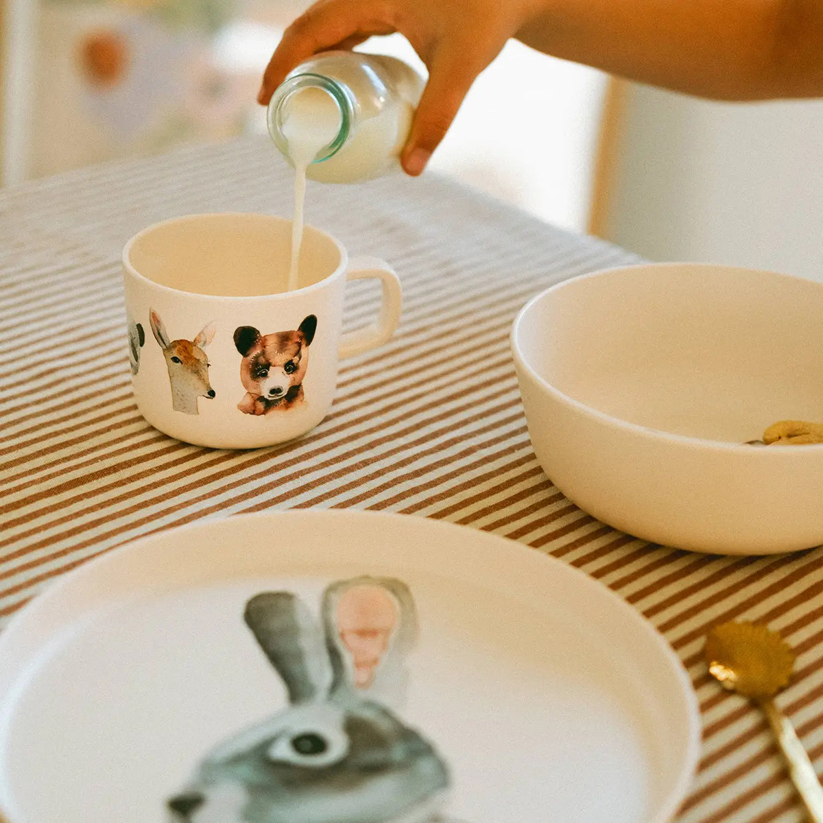 A hand pouring milk from a small glass bottle into a white ceramic mug decorated with animal faces, set on a striped tablecloth with a white plate and bowl nearby.
