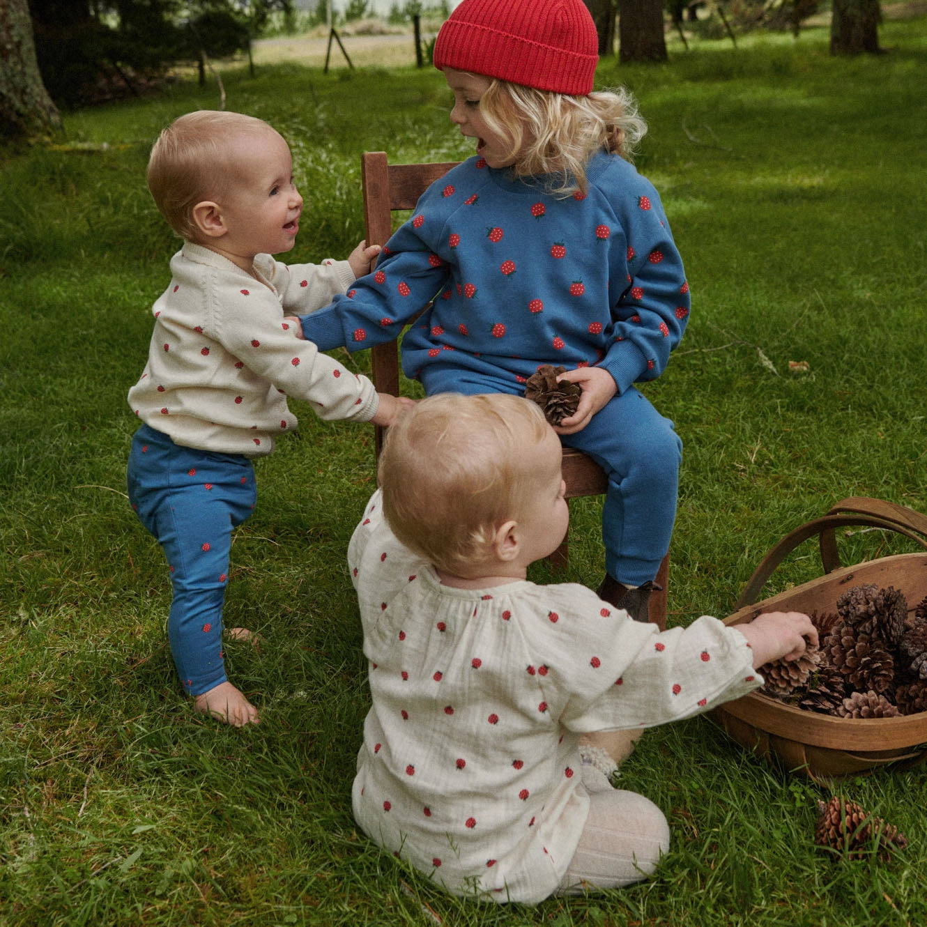 Three toddlers wearing matching cream and blue outfits with red polka dots play outdoors on a grassy lawn. One child sits on a wooden chair wearing a red knit beanie, while another stands and a third kneels near a basket of pinecones.