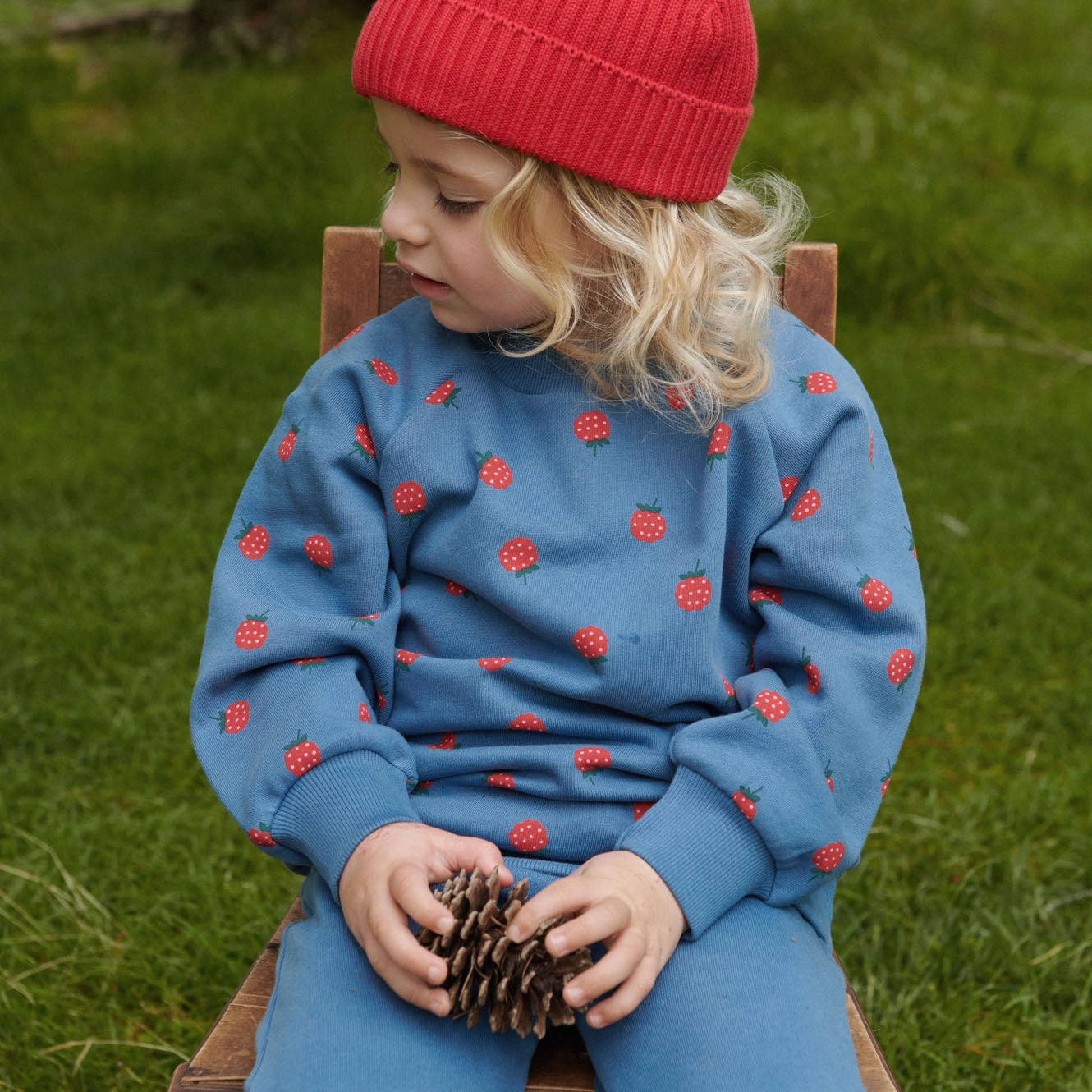 Child wearing a blue outfit with red patterns, sitting on a wooden chair outdoors.