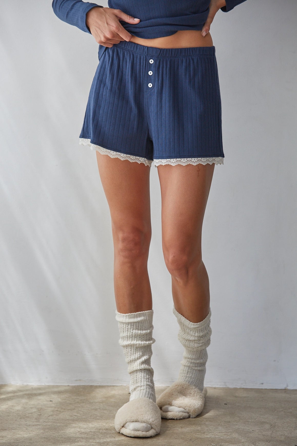 A person wearing blue ribbed shorts with white lace trim and a three-button front, paired with cream ribbed socks and slippers, standing against a light gray wall.