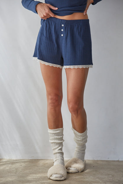 A person wearing blue ribbed shorts with white lace trim and a three-button front, paired with cream ribbed socks and slippers, standing against a light gray wall.
