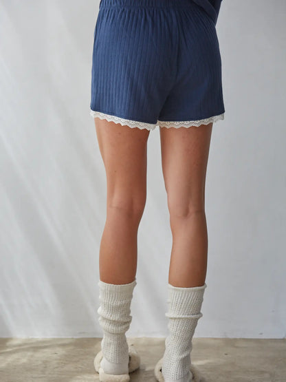 A person shown from the back wearing blue ribbed shorts with a white lace hem, paired with off-white ribbed socks and light-colored slippers against a plain light gray wall.