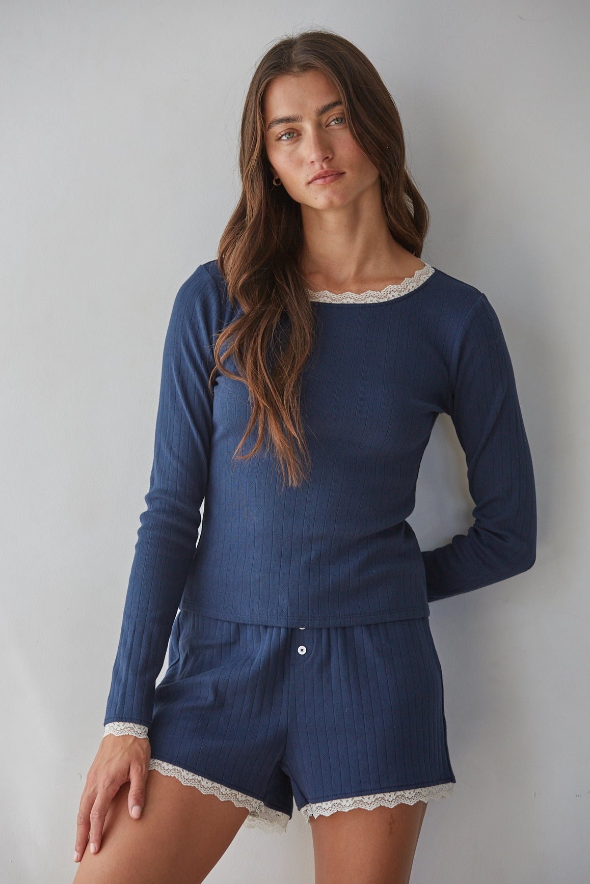A woman modeling a navy blue ribbed two-piece pajama set with white lace trim, consisting of a long-sleeve top and matching shorts, against a plain light gray background.