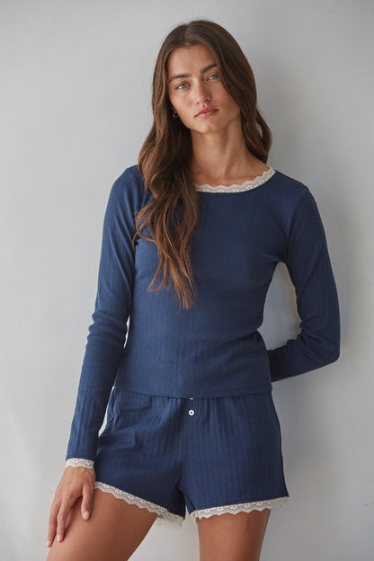 A woman modeling a navy blue ribbed two-piece pajama set with white lace trim, consisting of a long-sleeve top and matching shorts, against a plain light gray background.