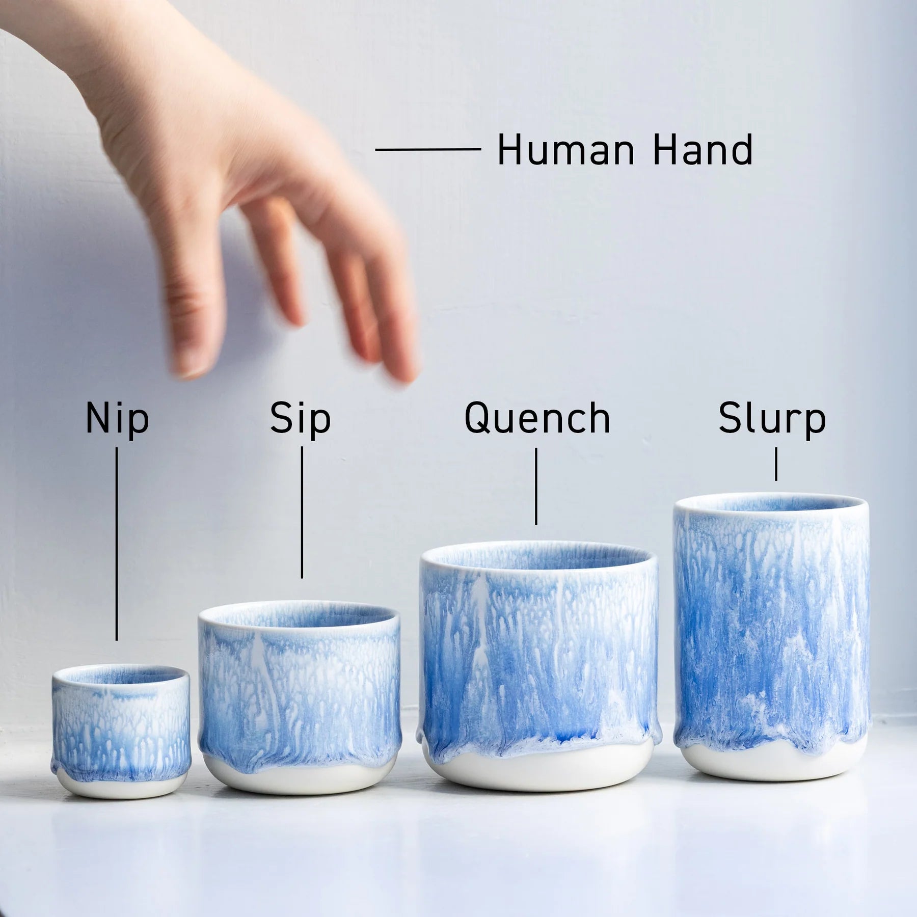 Set of four blue ceramic cups labeled 'Nip', 'Sip', 'Quench', and 'Slurp' with a hand reaching towards them. Studio Arhoj