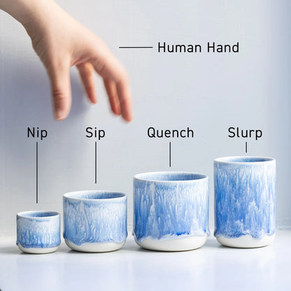 Set of four blue ceramic cups labeled 'Nip', 'Sip', 'Quench', and 'Slurp' with a hand reaching towards them. Studio Arhoj