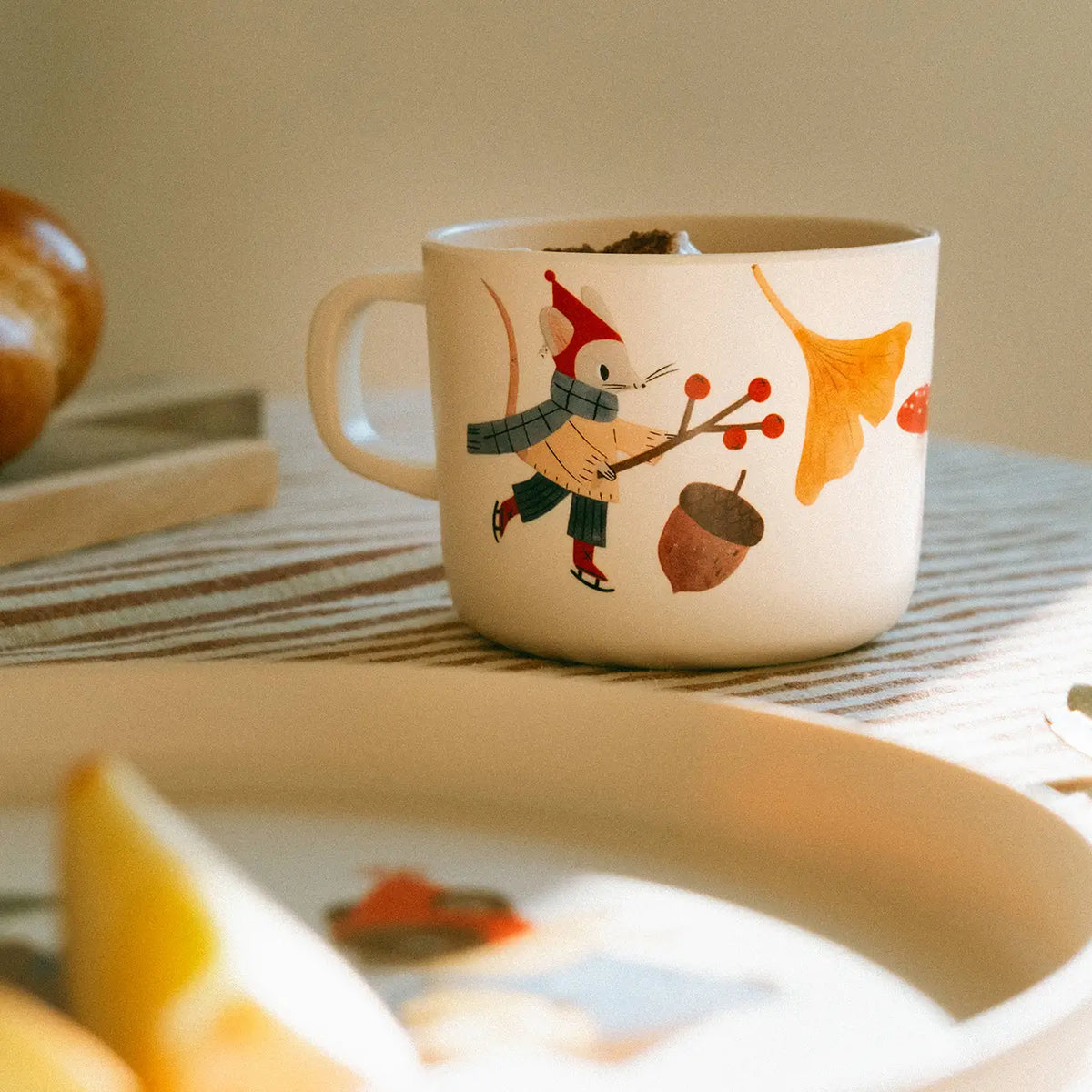 A cream-colored ceramic mug with a whimsical illustration of a mouse in a red hat and gray scarf holding a branch with red berries, alongside an acorn and orange ginkgo leaves. The mug is set on a striped table runner with a warm, softly blurred background.